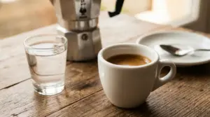 Tazzina di caffè espresso con bicchiere d’acqua su tavolo in legno, accanto a una moka e un piattino