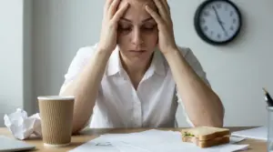 Persona con le mani sulla testa alla scrivania, con documenti e orologio sullo sfondo, segni di stress