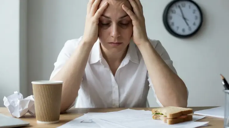 Persona con le mani sulla testa alla scrivania, con documenti e orologio sullo sfondo, segni di stress