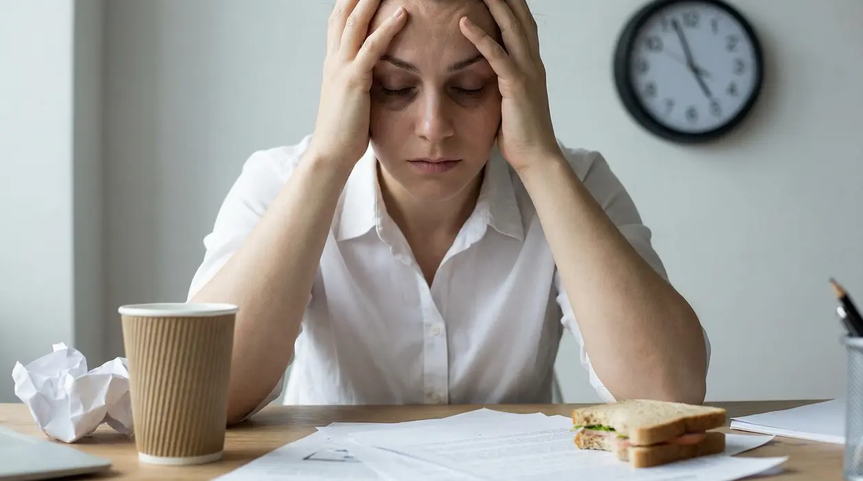 Persona con le mani sulla testa alla scrivania, con documenti e orologio sullo sfondo, segni di stress
