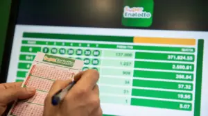 Mani che compilano una schedina del SuperEnalotto davanti a uno schermo con risultati e quote della vincita