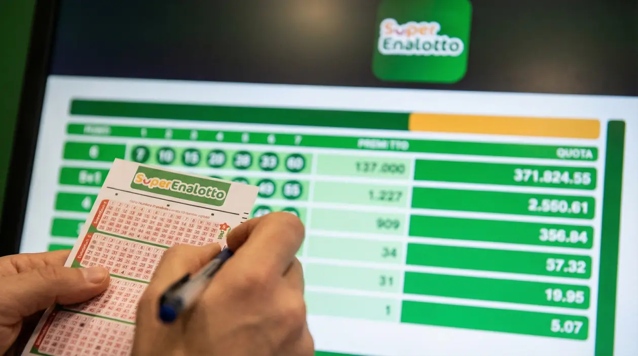 Mani che compilano una schedina del SuperEnalotto davanti a uno schermo con risultati e quote della vincita